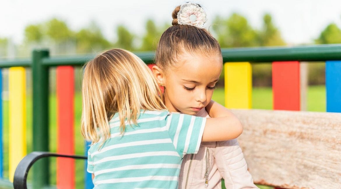 Two Young Girls Hugging On A Bench In A Park.
